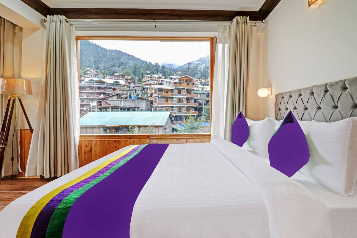 Habitación de hotel con cama, cortinas, lámpara y vistas a un pueblo montañoso.