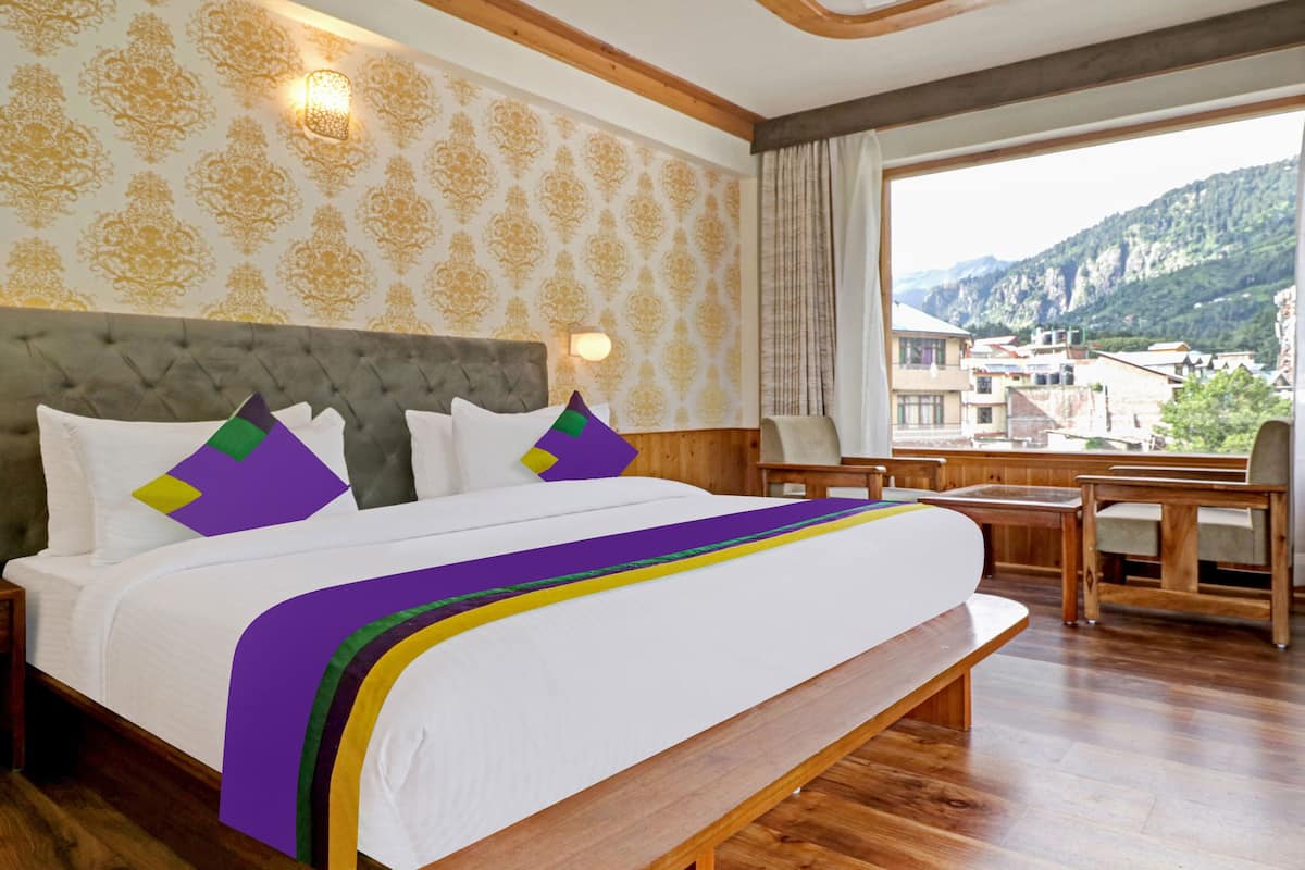 Habitación de hotel con una cama grande, cabecera de madera, vistas a la montaña y un balcón con mobiliario de madera.