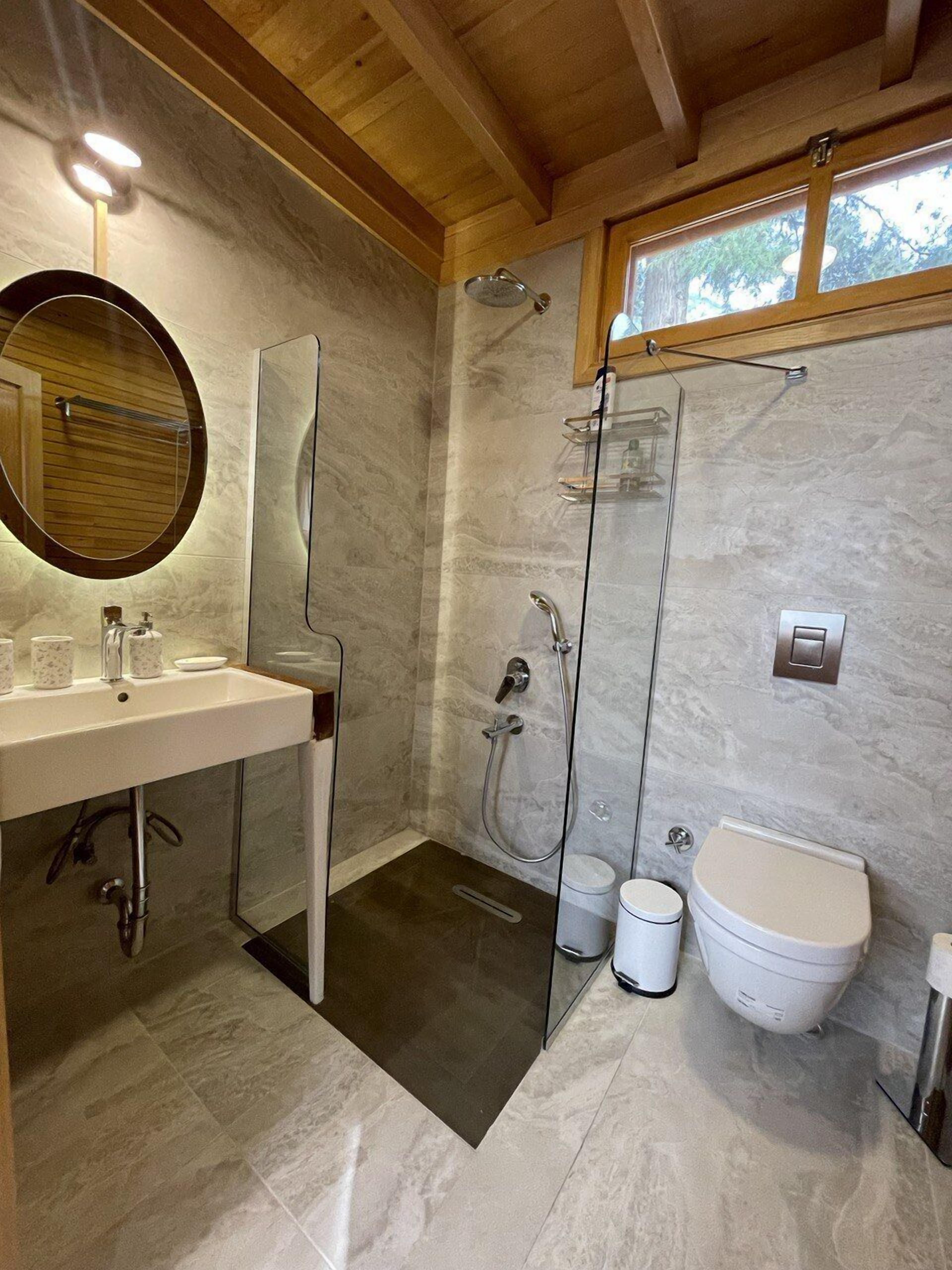 A modern bathroom with a marble floor, a wooden ceiling, a round mirror, a white sink, a glass shower enclosure, and a toilet.