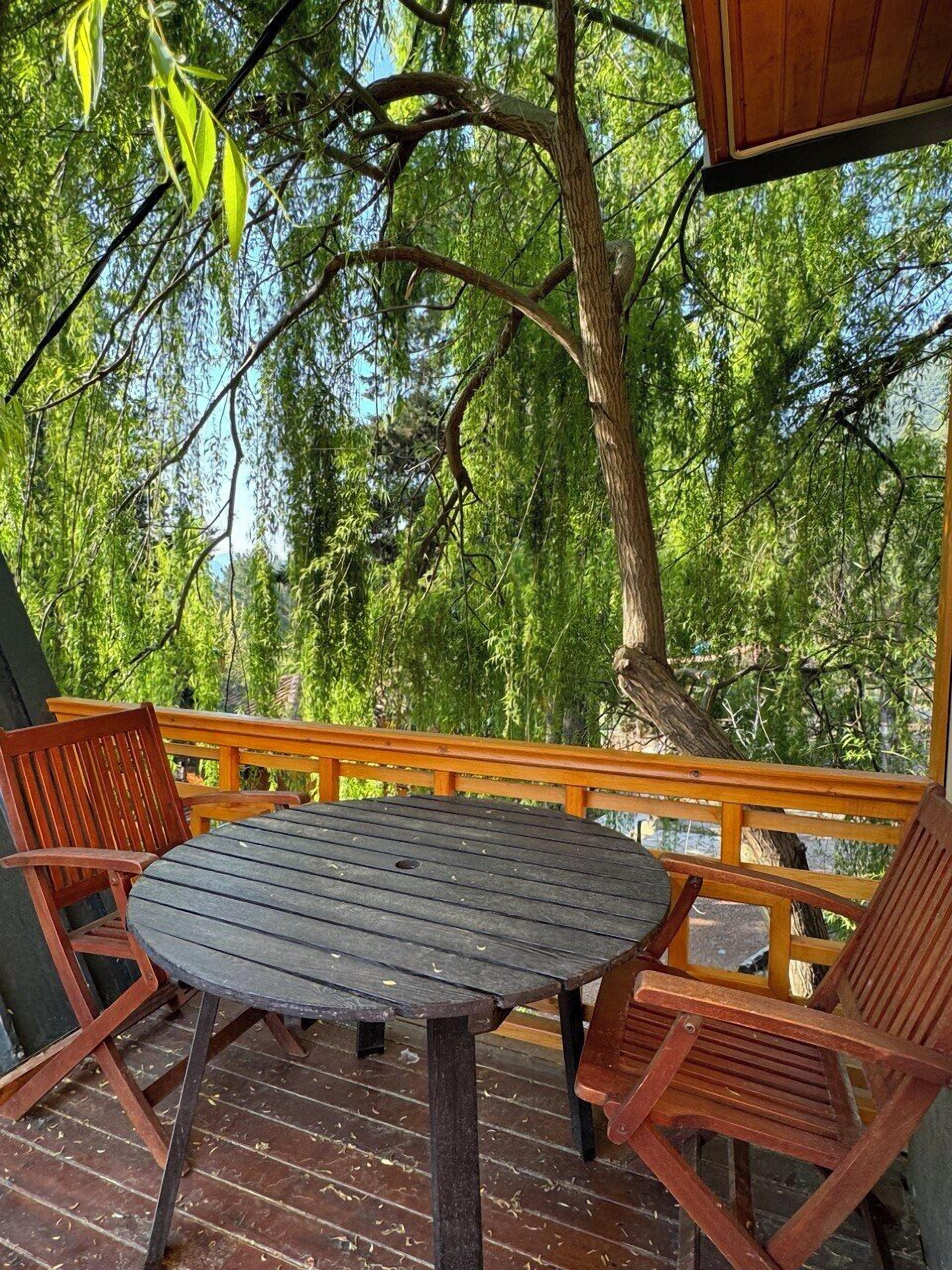 Una terraza de madera con una mesa redonda y dos sillas, rodeada de árboles verdes y frondosos.