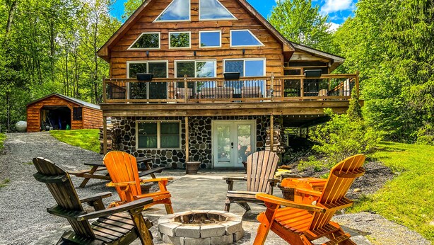 A log cabin with a stone foundation, a balcony, and a fire pit surrounded by Adirondack chairs.