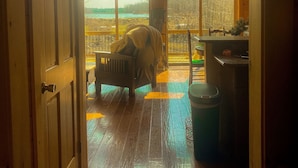 A room with a wooden floor, a bed, a chair, and a table. There are large windows providing a view of the outdoors.