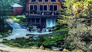 A two-story cabin with a stone foundation, a balcony with outdoor seating, and a surrounding natural landscape.