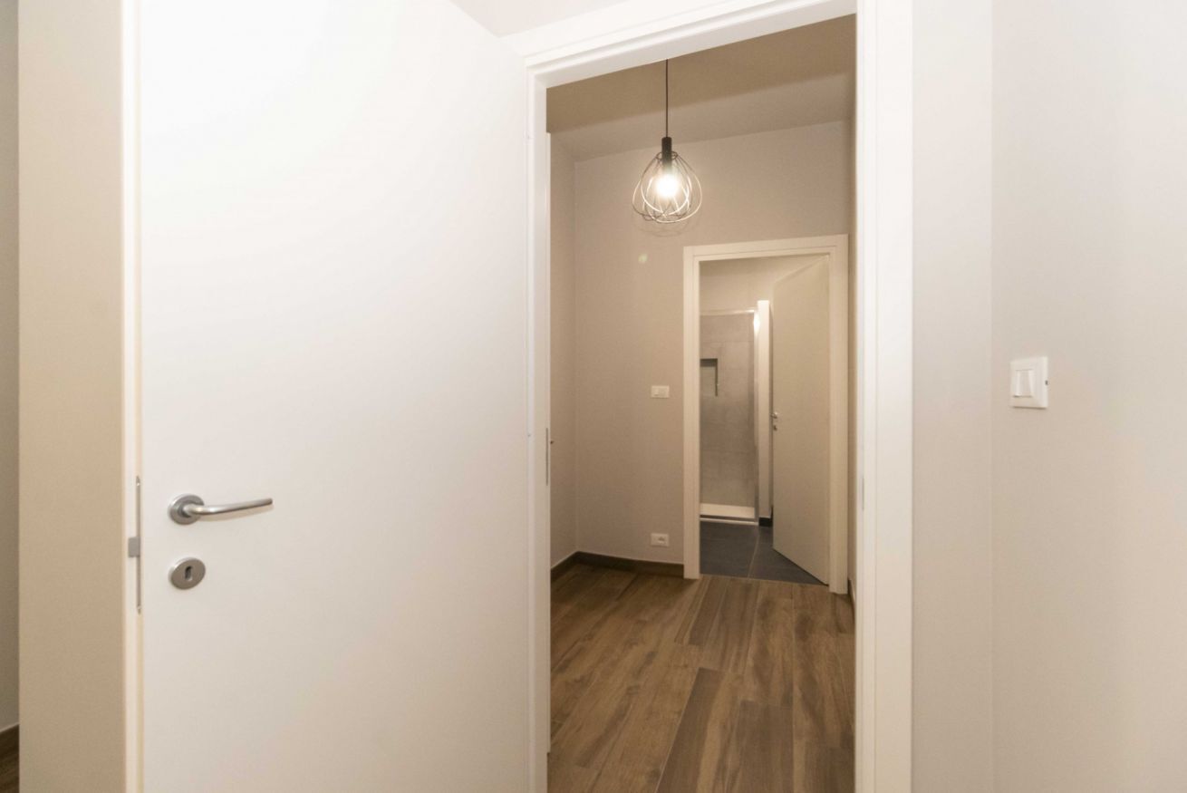 A hallway with a wooden floor, a white door with a silver handle, and a hanging light fixture.