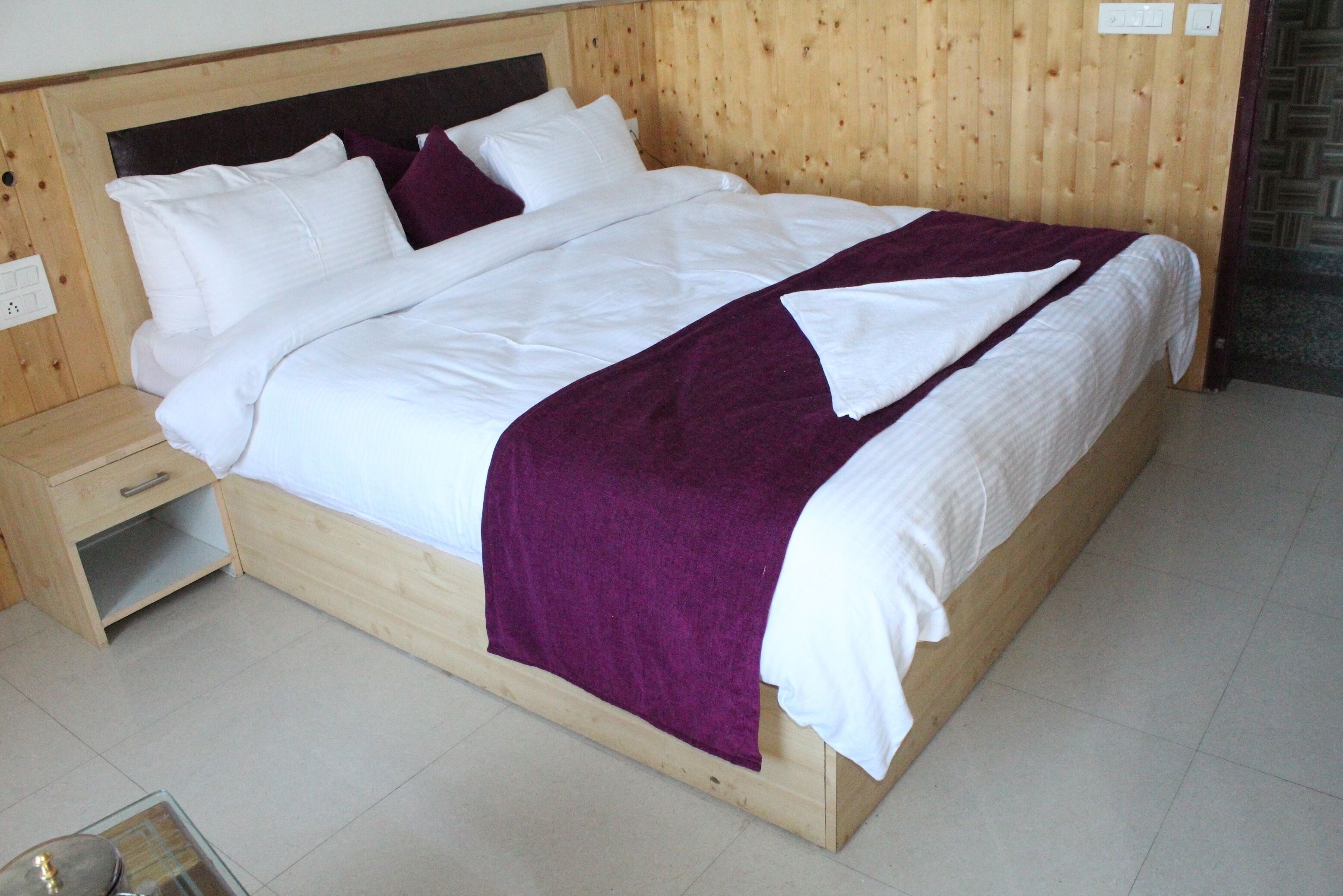 Een net opgemaakt bed met wit en paars beddengoed, een houten hoofdbord en een klein nachtkastje met lade.