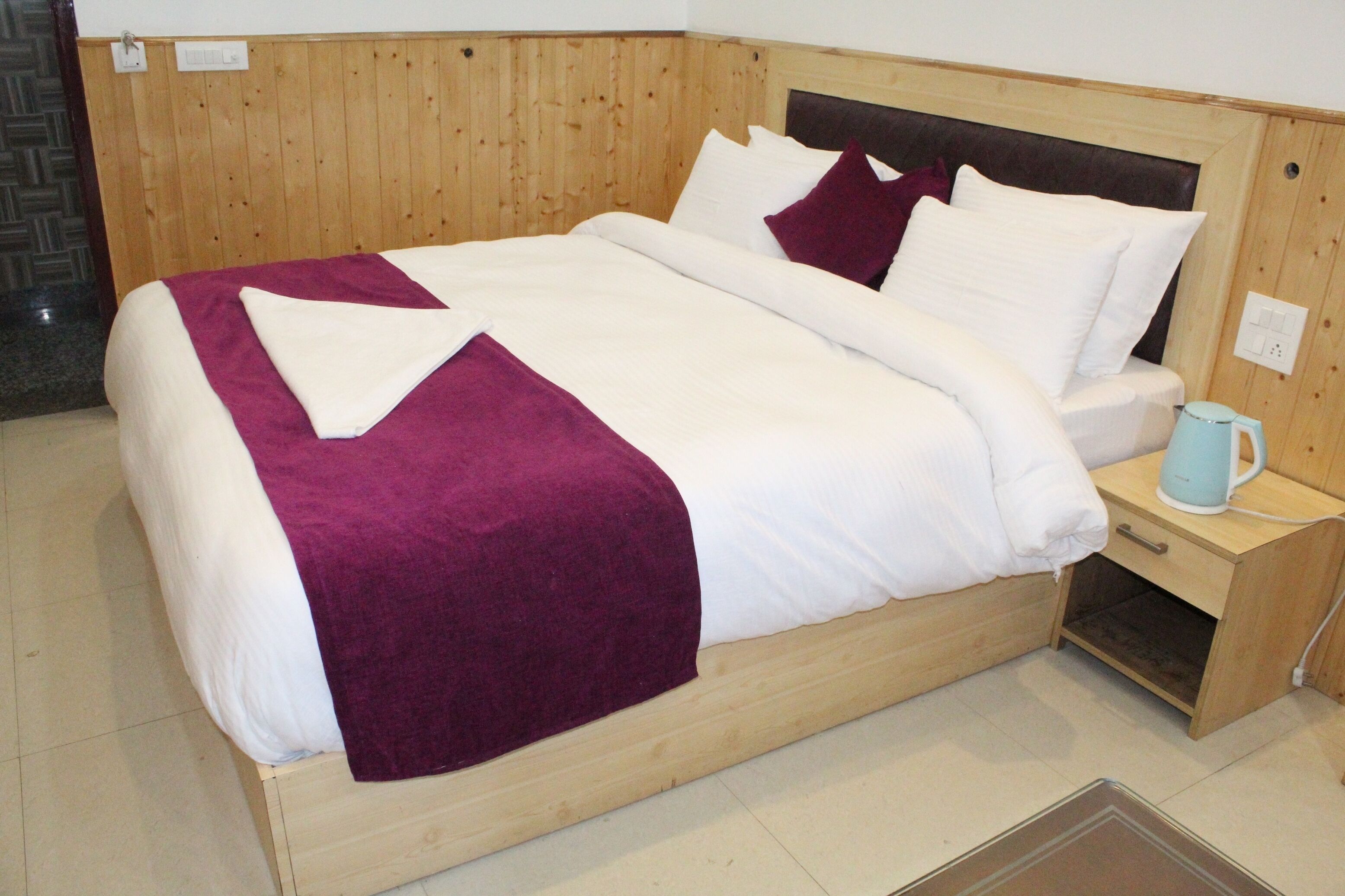 A wooden bed with white and purple bedding, a nightstand with a blue mug, and a tiled floor.