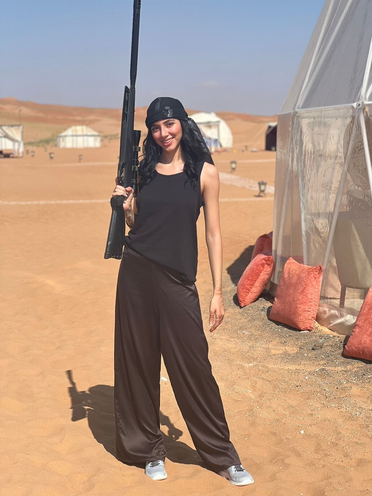 A person standing in a desert holding a rifle, with tents and cushions in the background.
