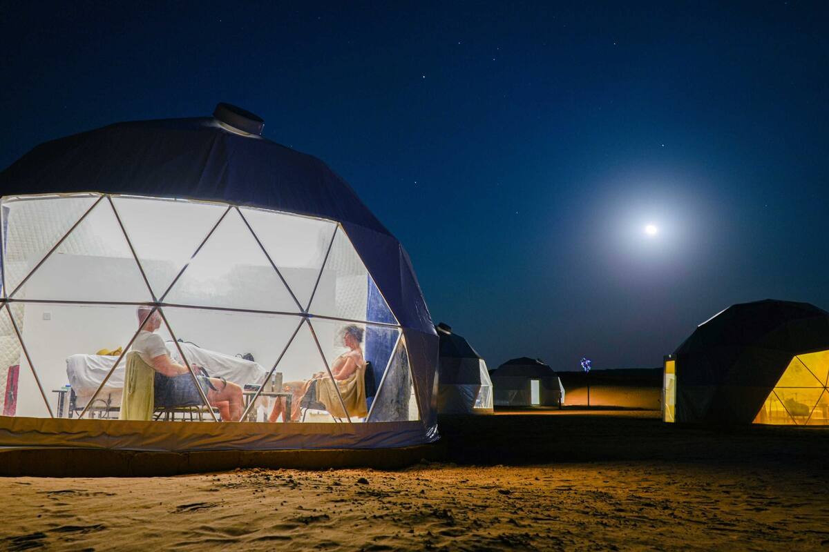 A series of geodesic domes with a clear view of the interior, including a dining area with tables and chairs, set against a night sky with a visible moon.
