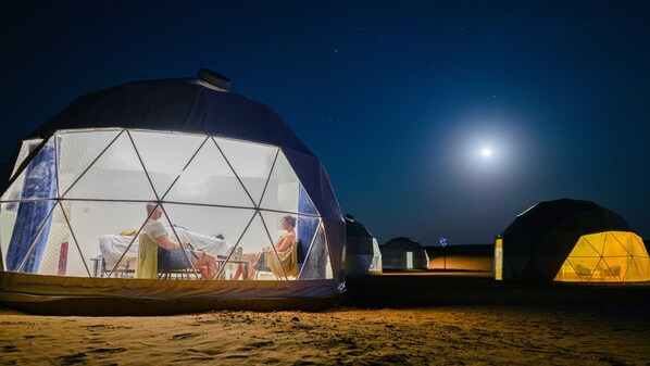 Una serie de cúpulas geodésicas con vista despejada al interior, incluyendo un comedor con mesas y sillas, frente a un cielo nocturno con luna visible.