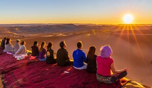 A group of people sitting on a red carpeted area outdoors, overlooking a desert landscape with sand dunes and a sunset. - Luxury Desert Camp Oman (Bidiya)