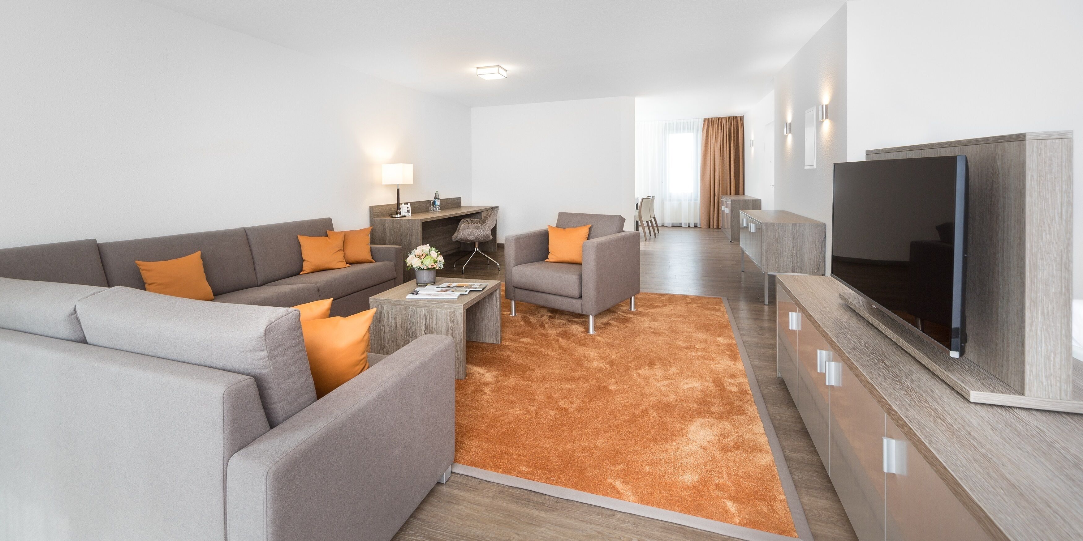 Ein modernes Wohnzimmer mit grauem Sofa, orangefarbenen Kissen, einem Couchtisch, einem Fernseher und einer Wand aus Holz.