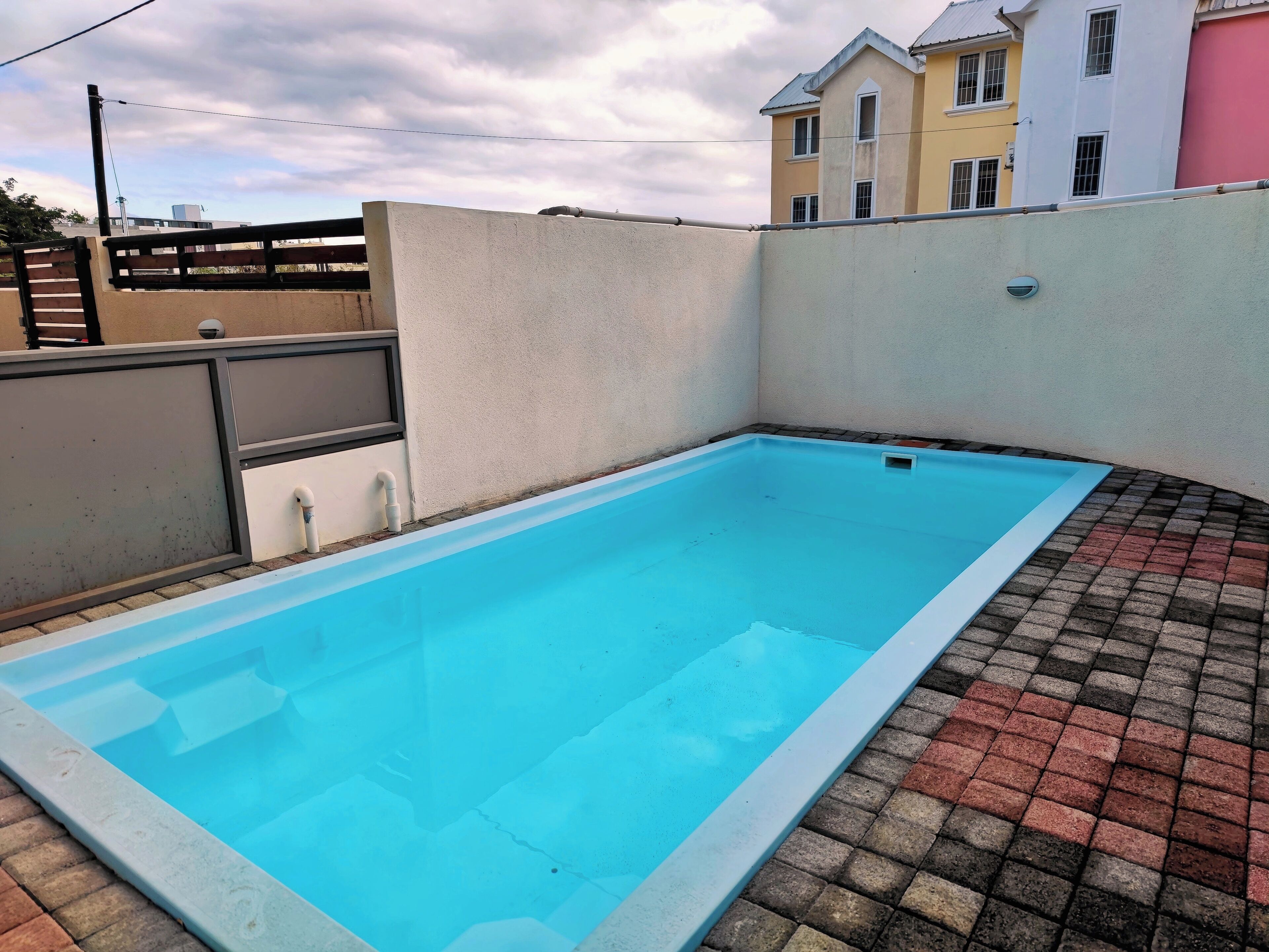 Una piscina rectangular con borde alicatado, rodeada por un muro bajo y un área pavimentada con ladrillos rojos y grises.
