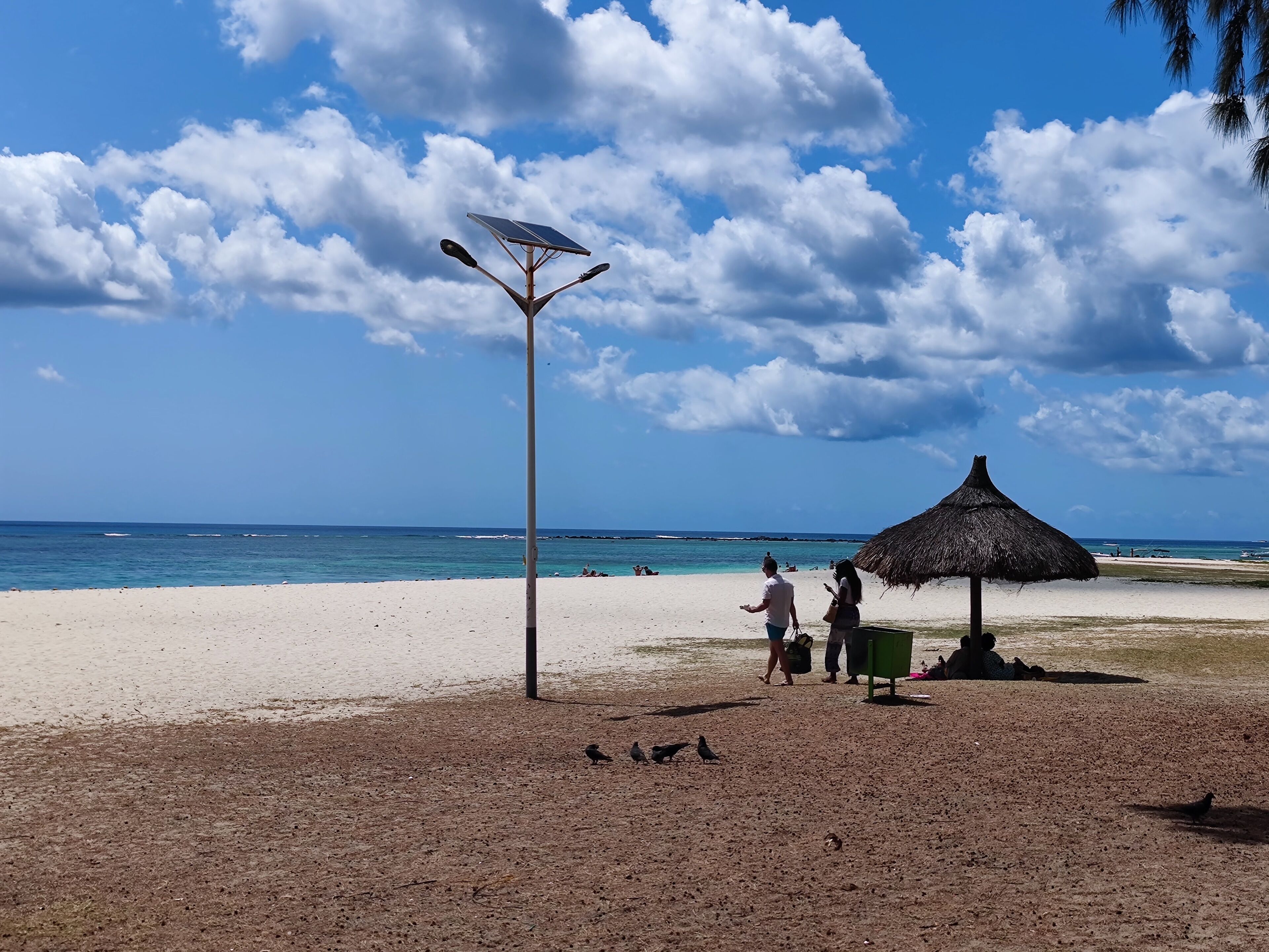 A beach with a thatched umbrella and a solar-powered street lamp.