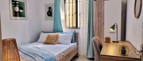 A bedroom with a bed, a desk, a chair, a lamp, a hanging basket, and a window with curtains.