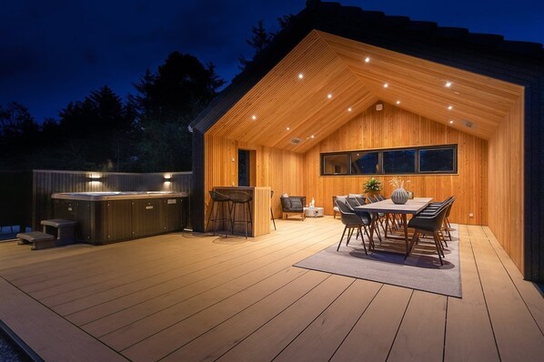 A modern wooden cabin with a deck, a hot tub, and a dining area with chairs and a table.