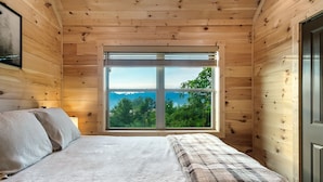 A wooden cabin bedroom with a large window, a bed with a plaid blanket, and a nightstand with a lamp. - Prime Location! Relaxing 2/2 Luxury Cabin! Views, Fire Pit, Hot Tub & Game Room. (Gatlinburg)