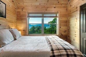 Ein Schlafzimmer einer Holzhütte mit einem großen Fenster, einem Bett mit karierter Decke und einem Nachttisch mit Lampe.