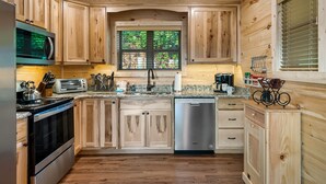 A modern kitchen with wooden cabinets, stainless steel appliances, granite countertops, and a window with a view of greenery. - Prime Location! Relaxing 2/2 Luxury Cabin! Views, Fire Pit, Hot Tub & Game Room. (Gatlinburg)