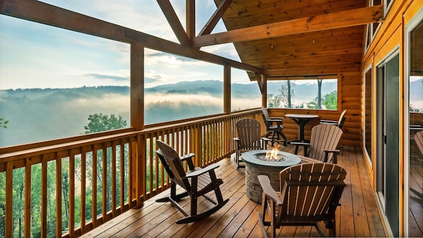 A wooden deck with Adirondack chairs and a fire pit, overlooking a forested valley and mist-covered mountains. - Prime Location! Relaxing 2/2 Luxury Cabin! Views, Fire Pit, Hot Tub & Game Room. (Gatlinburg)