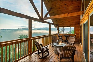 Um deck de madeira com cadeiras Adirondack e uma lareira, com vista para um vale arborizado e montanhas envoltas em névoa.