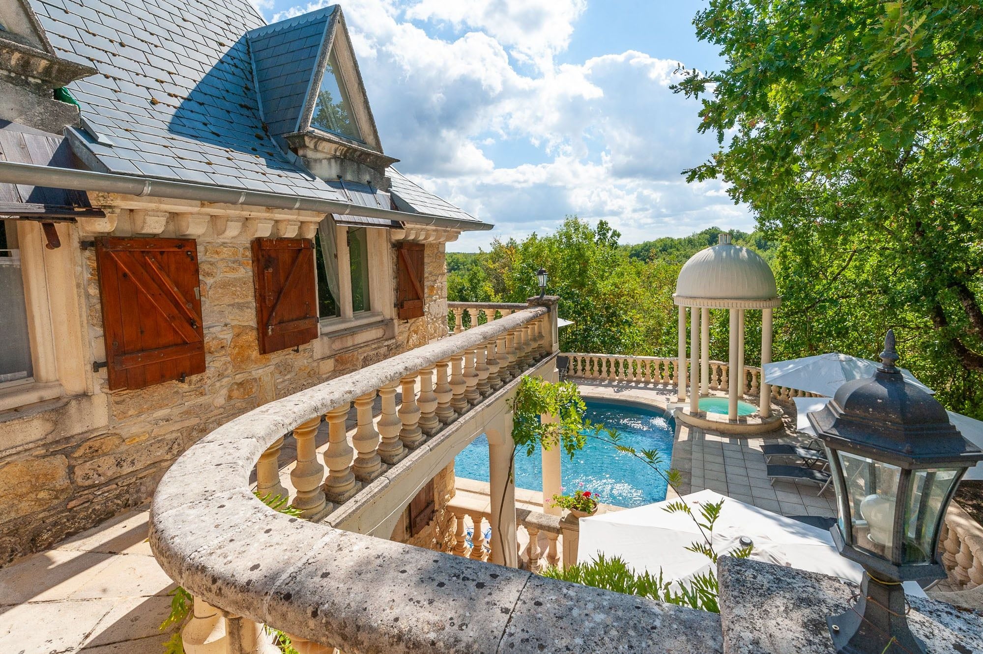 A stone building with a balcony overlooking a swimming pool, surrounded by lush greenery.
