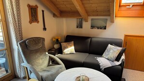 A cozy living room with a wooden ceiling, a grey armchair, a black sofa, and a small round table with a candle.