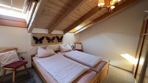 A cozy attic bedroom with a wooden ceiling, two beds with matching headboards, a chair, and a wall-mounted lamp.