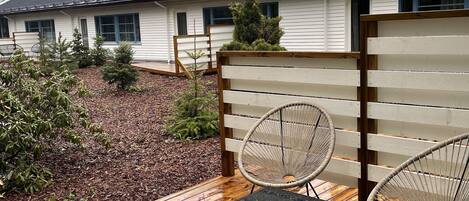 A wooden deck with two wicker chairs and a small table, surrounded by a garden with mulch and small plants.