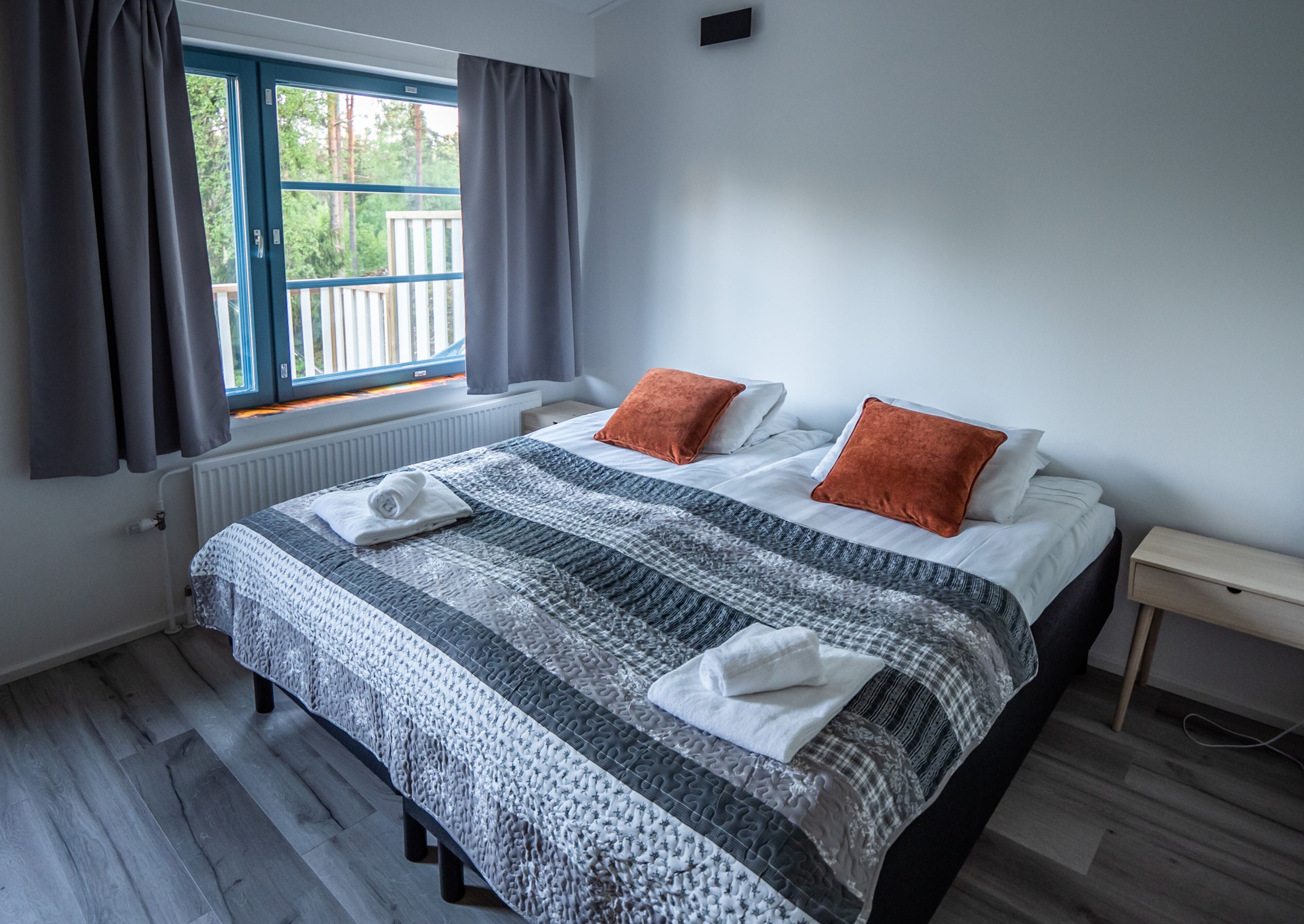 Quarto com cama grande, cortinas cinzas, janela com vista para árvores e piso de madeira.