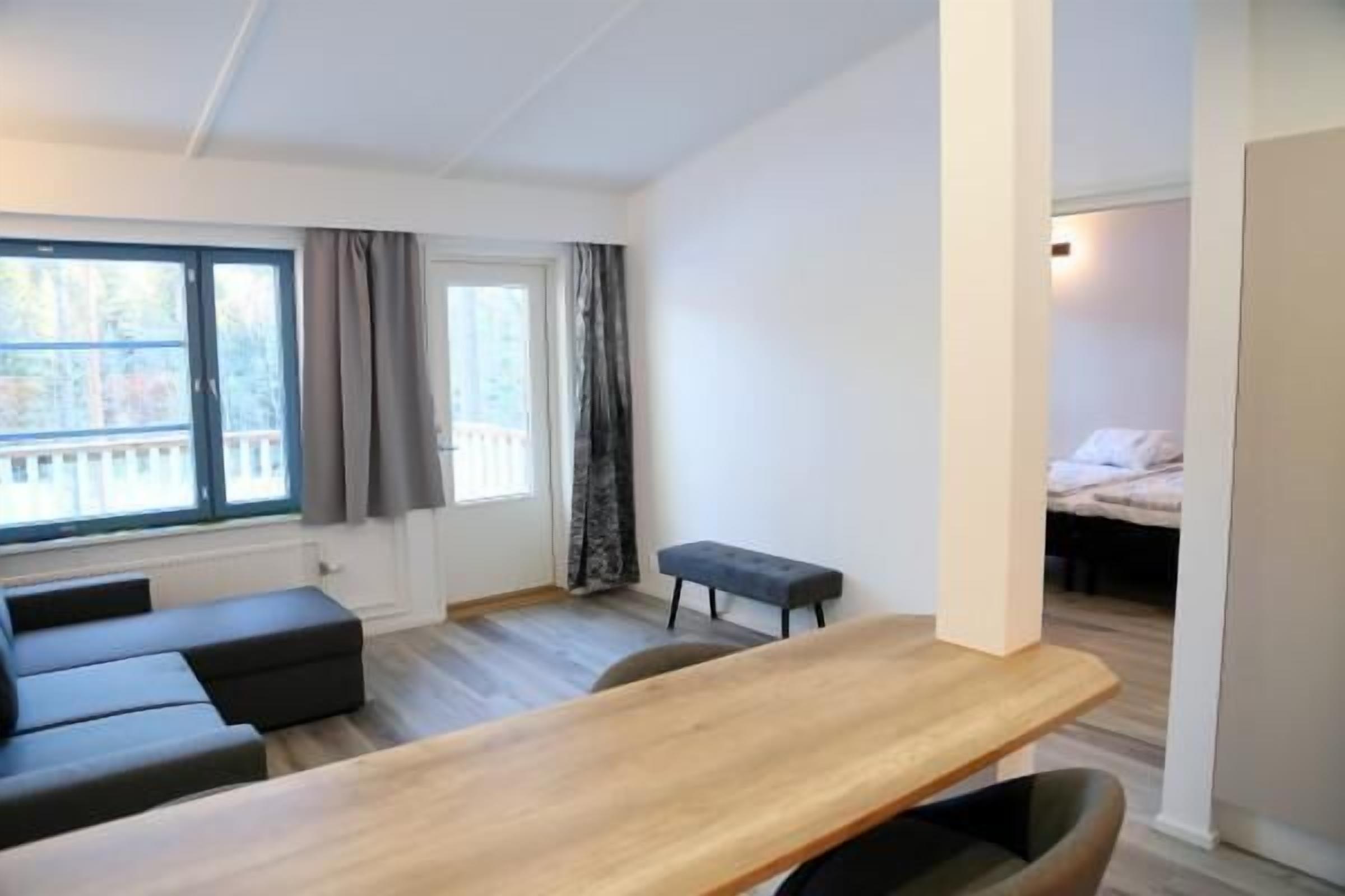 Una habitación moderna con una mesa de madera, un sofá negro y un pequeño banco. Hay una ventana con cortinas y vistas a árboles.