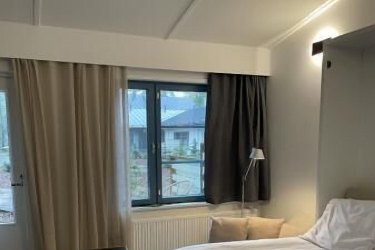 A hotel room with two beds, a window with curtains, a radiator, a bedside table with a lamp, and a view of the outside.