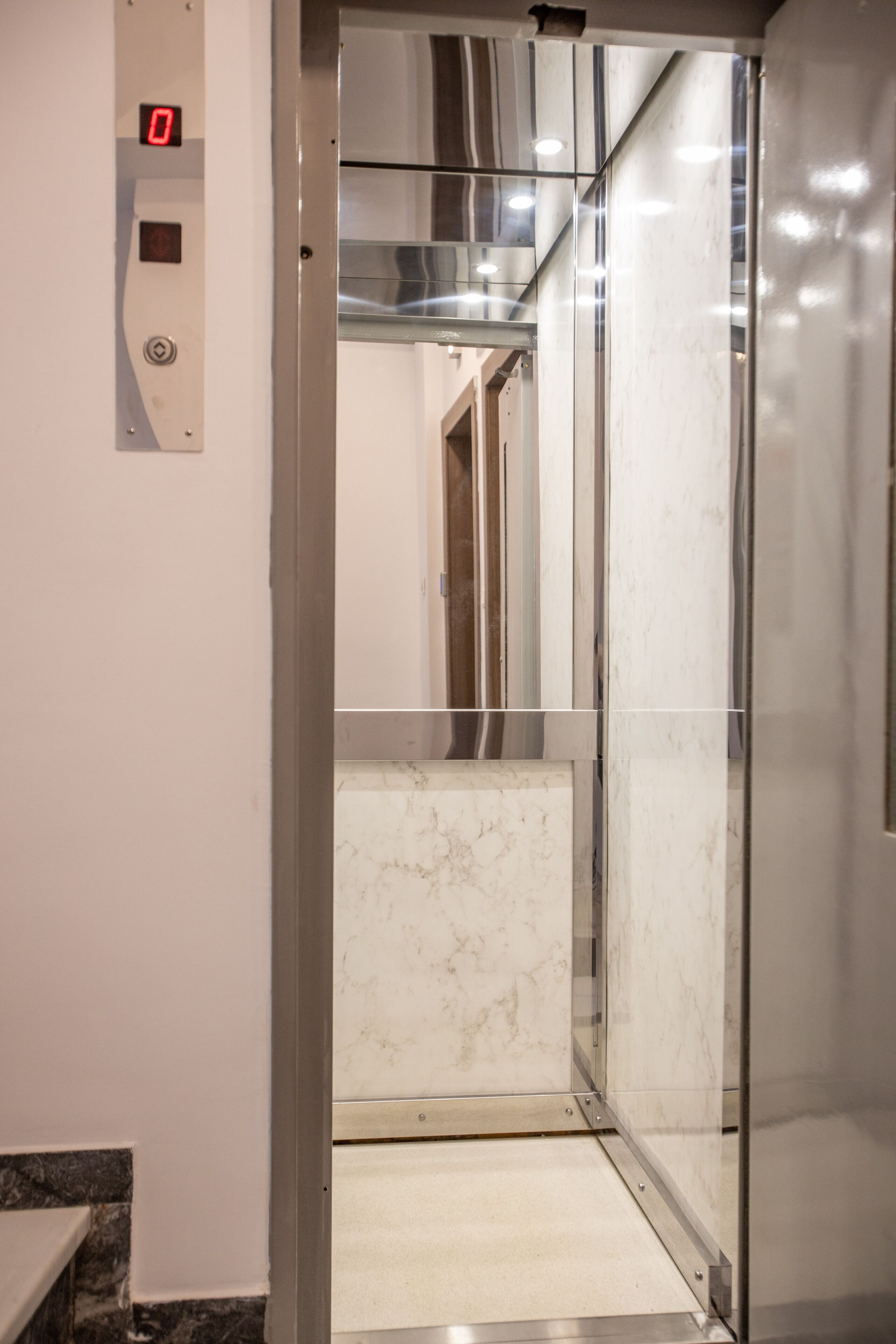 Interior de elevador com painel digital, paredes de mármore e chão metálico.