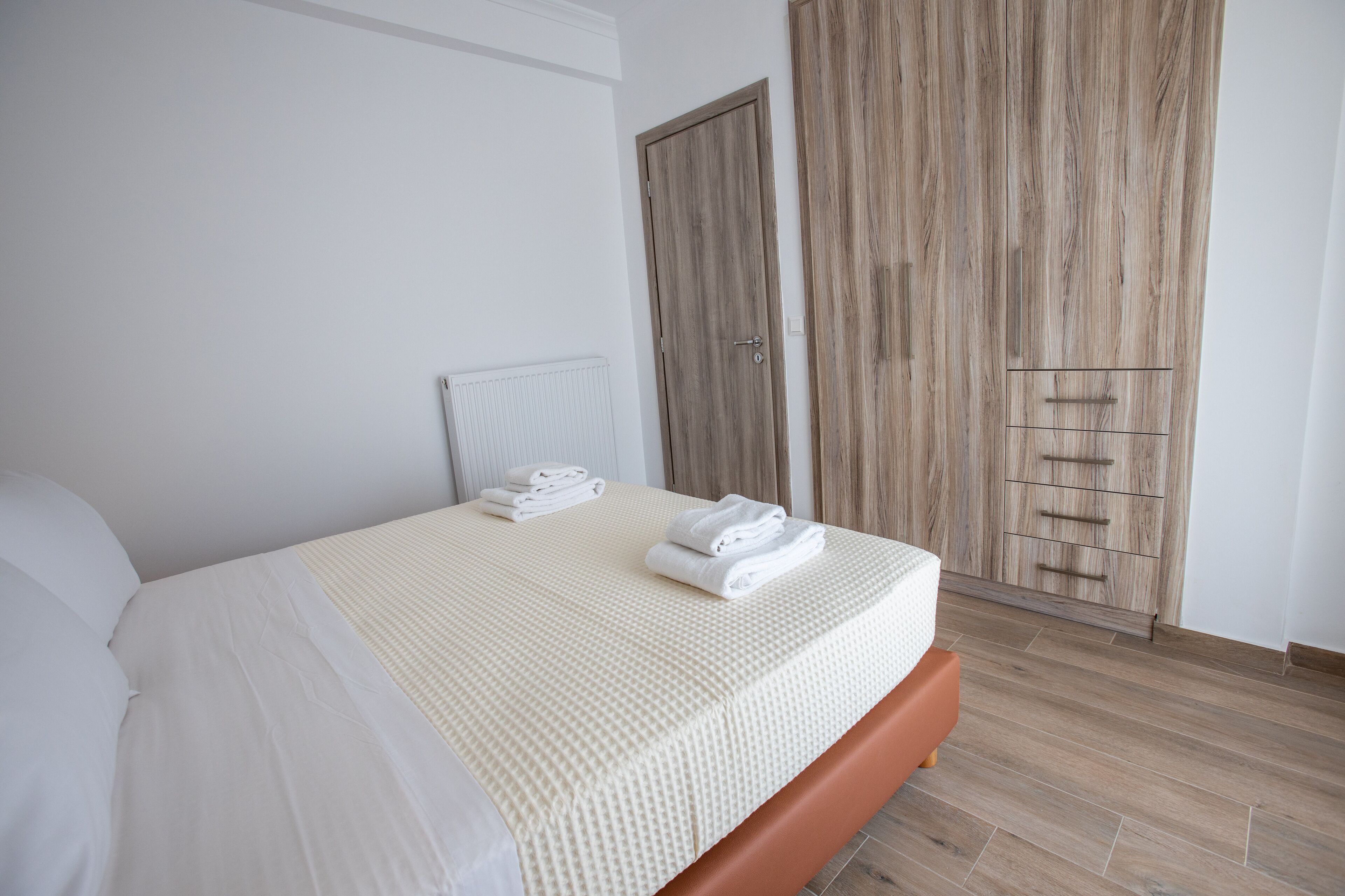A neatly made bed with white linens and a beige blanket, a wooden wardrobe with a mirror, and a radiator on the wall.