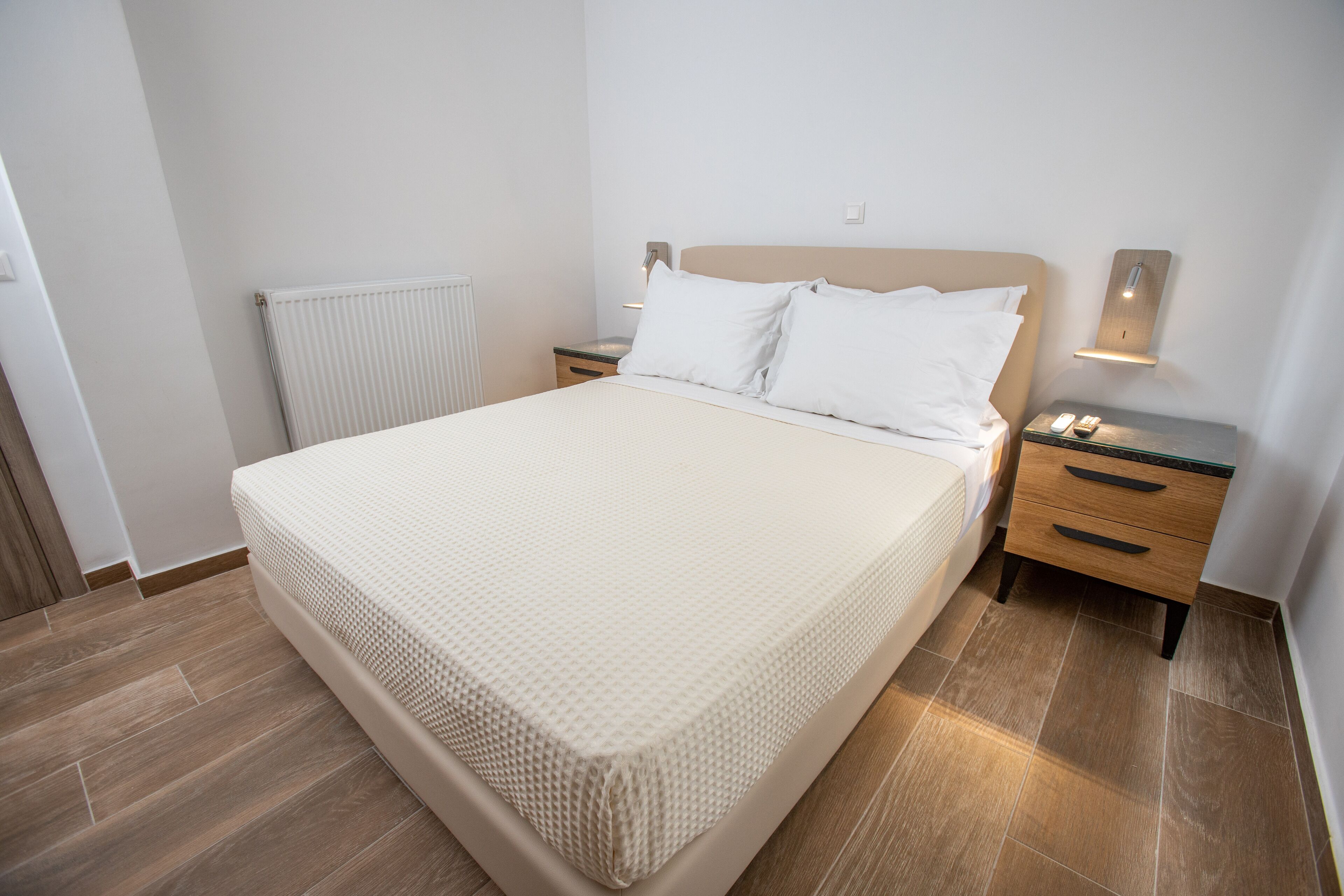 A neatly made bed with a white mattress and pillows, a wooden nightstand with a lamp, and a radiator on the wall.