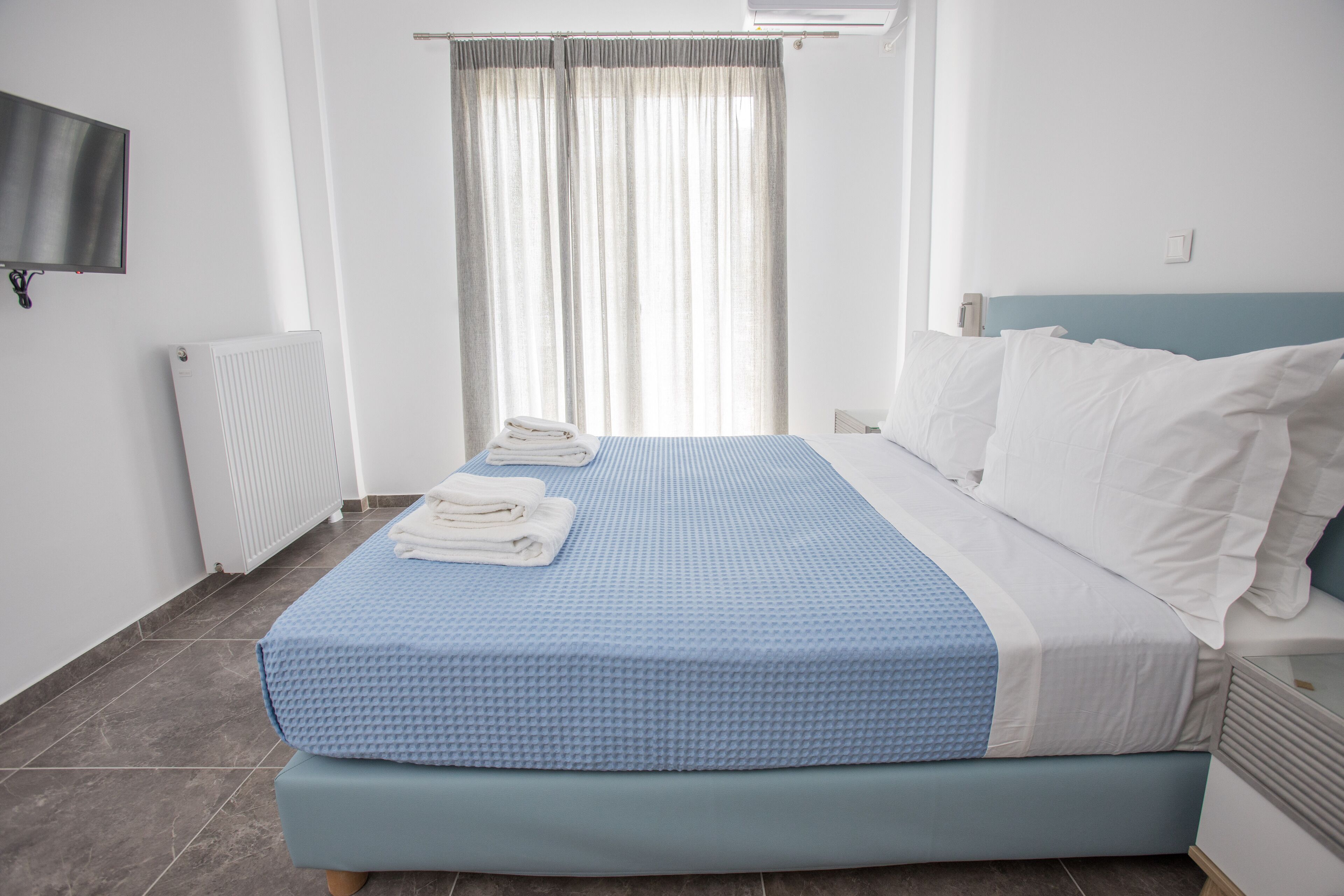 A hotel room with a bed, a TV, a radiator, and a window with curtains.
