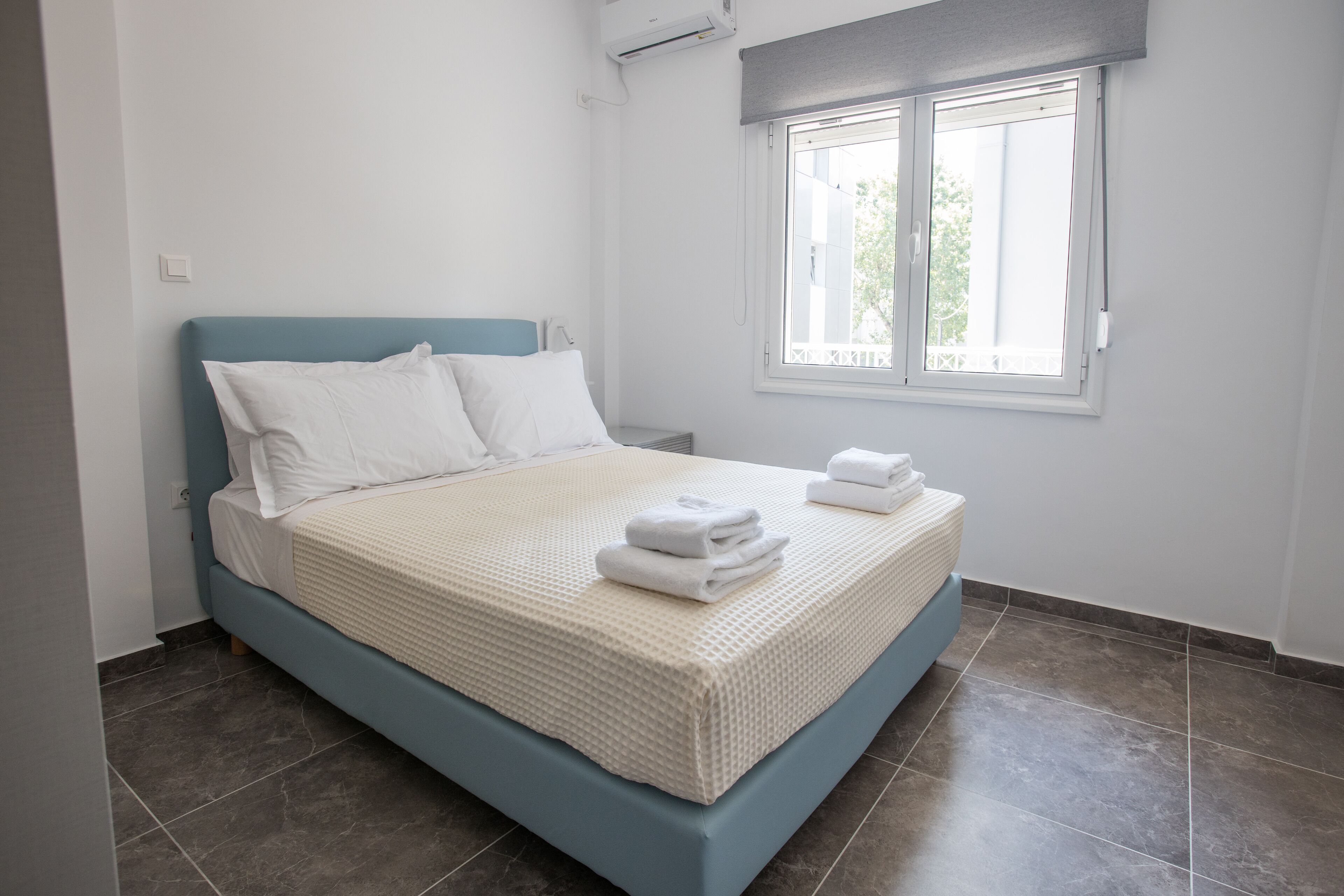 Una cama bien hecha con sábanas blancas y un cabecero azul en una habitación con un ventanal grande y suelo de baldosas.