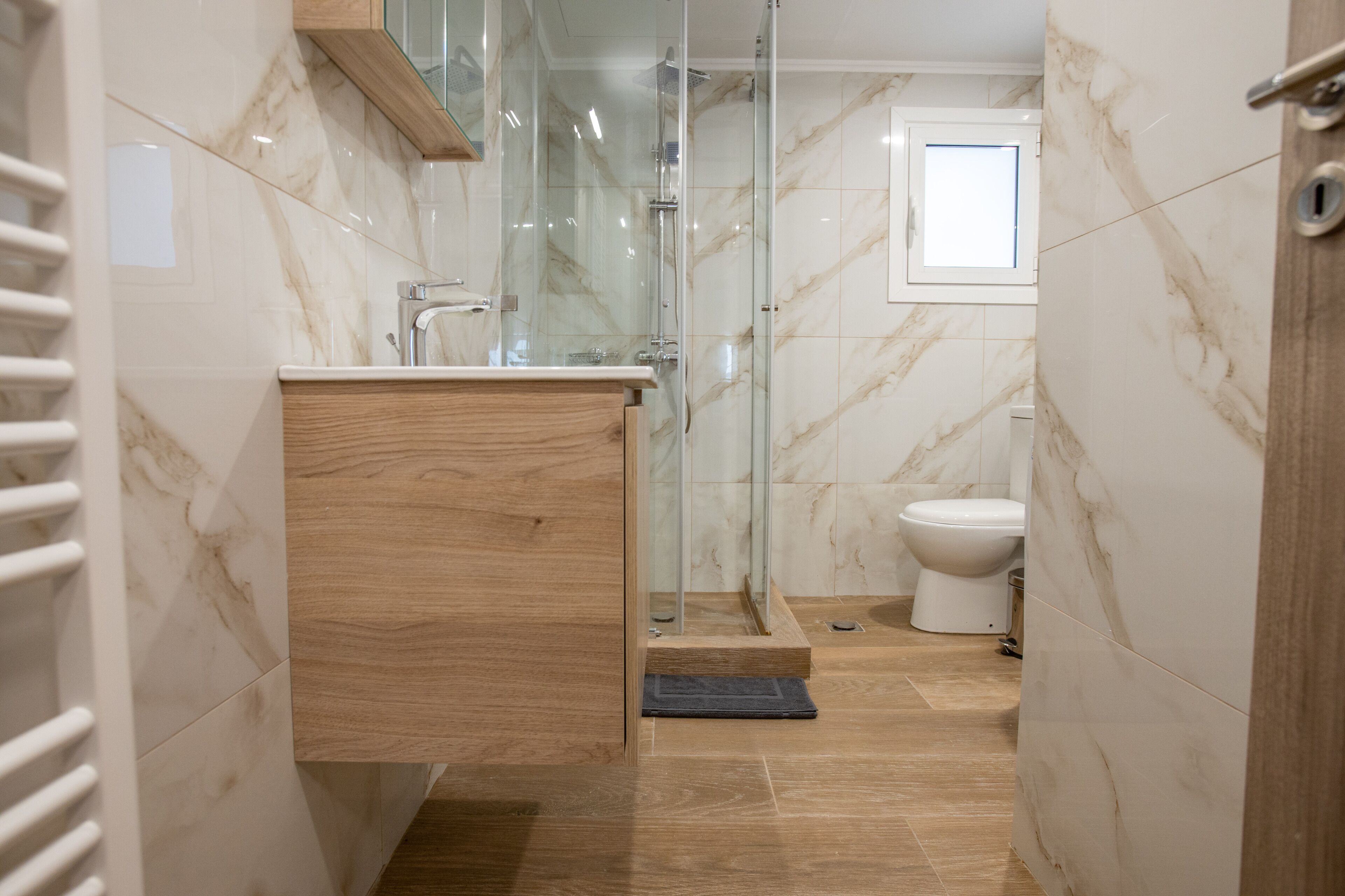 A modern bathroom with a glass shower enclosure, a wooden vanity, and a marble-tiled floor.