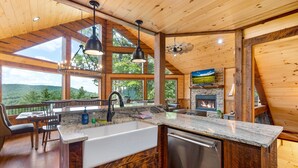 A kitchen with a stone fireplace, wooden cabinets, and a large window overlooking a forested area. - Tyson’s Peak – Blue Ridge, GA. (Blue Ridge)