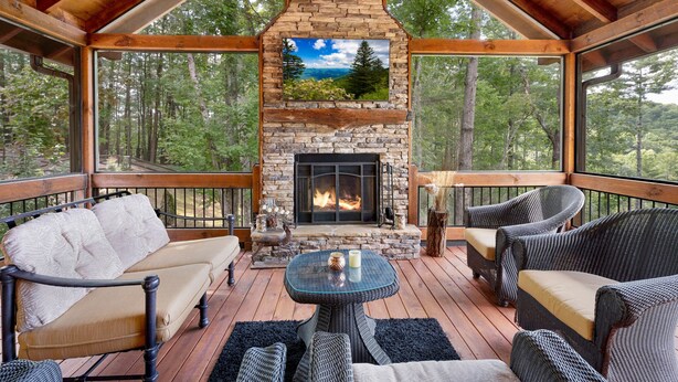 A covered deck with a stone fireplace, wicker furniture, and a scenic view of trees. - Tyson’s Peak – Blue Ridge, GA. (Blue Ridge)