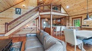A spacious loft-style room with a high ceiling, wooden beams, a large leather sofa, a dining table with chairs, a kitchen area, and a staircase leading to an upper level. - Tyson’s Peak – Blue Ridge, GA. (Blue Ridge)