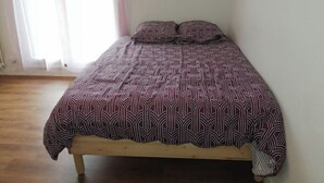 A single bed with a patterned bedspread and pillows in a room with wooden flooring and a window with sheer curtains. - Studio Bienvenue Tout Confort (Orléans)