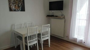 A room with a white table and chairs, a painting of a leopard, a flat-screen TV mounted on the wall, and a window with curtains. - Studio Bienvenue Tout Confort (Orléans)