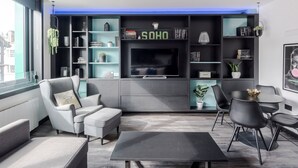 A modern living room with a grey sofa, a black coffee table, and a TV on a black stand. There are bookshelves with books and decorative items, and a window with a view of buildings. - West End 2-Bed · Soho · Sleeps 6 (London)