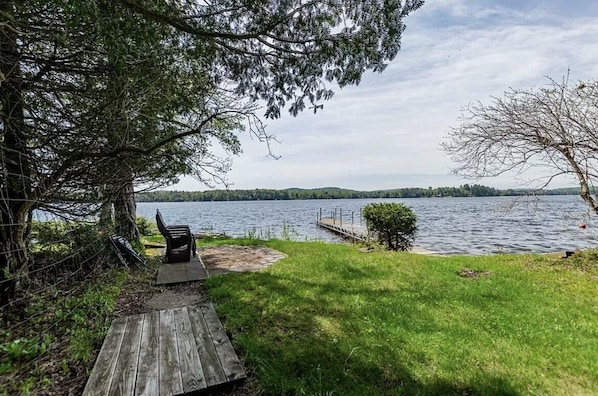A lakeside deck with a wooden bench and a small table, surrounded by lush greenery and a clear view of the lake.