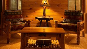 Interior acogedor de una cabaña con paredes de madera, un ventilador de techo, un cuadro enmarcado, una mesa con lámpara y dos sillones estampados.