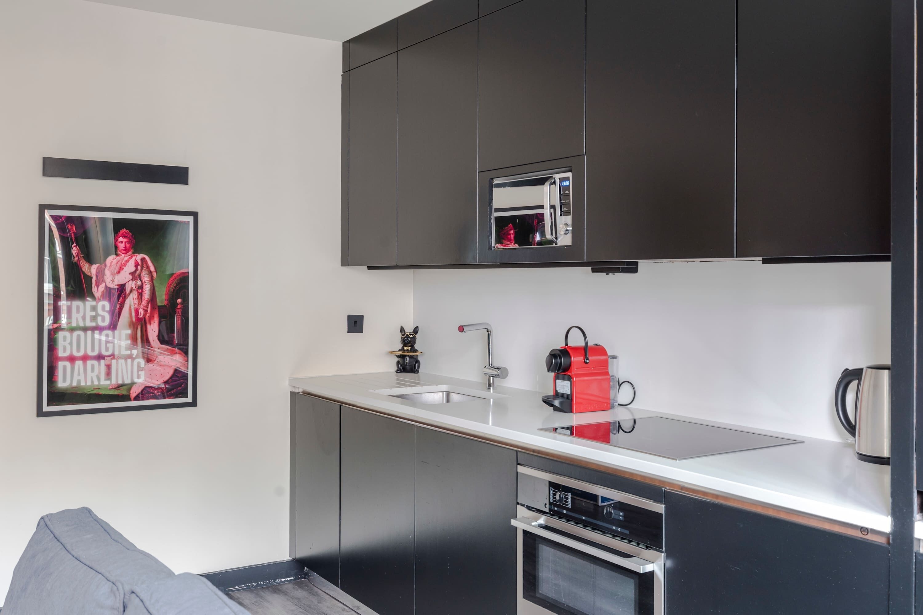 Una cocina moderna con armarios oscuros, un lavamanos de acero inoxidable, un microondas integrado, un hervidor rojo y un cartel en la pared.