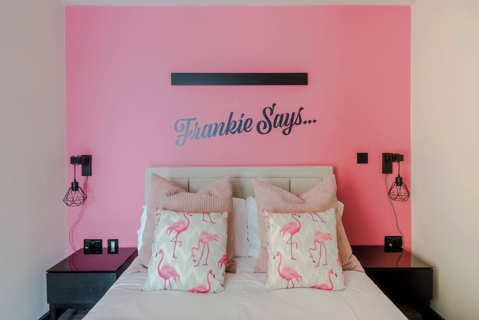 Ein Schlafzimmer mit rosa Wand, einem Bett mit weißer Bettwäsche und Kissen mit rosa Flamingos sowie einem schwarzen Regal mit einer Lampe und einem kleinen Tisch.