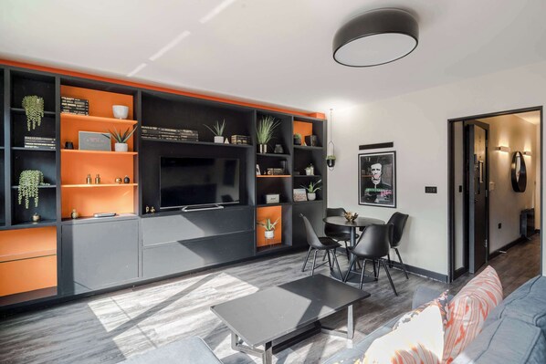 Una sala de estar moderna con un sofá, una mesa de centro y un comedor con sillas. Hay un televisor en un mueble negro para entretenimiento, y estantes con objetos decorativos y plantas.