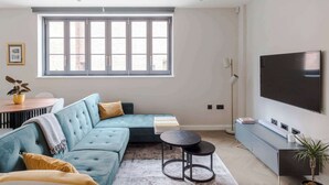 A modern living room with a sectional sofa, a round coffee table, a flat-screen TV, and a large window with blinds. - Stunning 2-Bed Mews House w/ AC near Regent's Park (London)