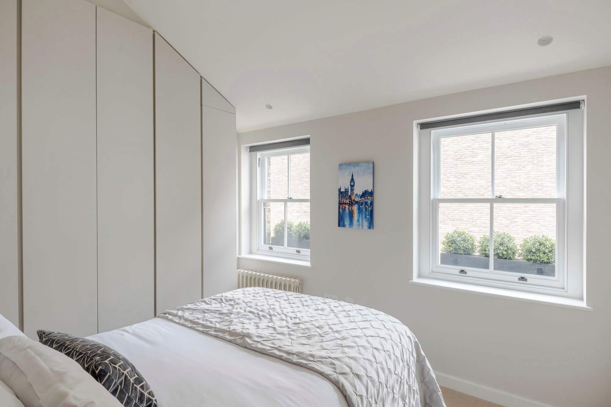 A bedroom with a bed, a radiator, and two windows with a cityscape painting.